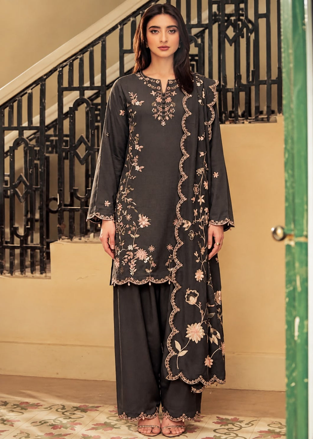 A woman modeling a dark charcoal gray three-piece kurta set with beige floral embroidery, standing in front of a beige wall and black metal staircase railing.