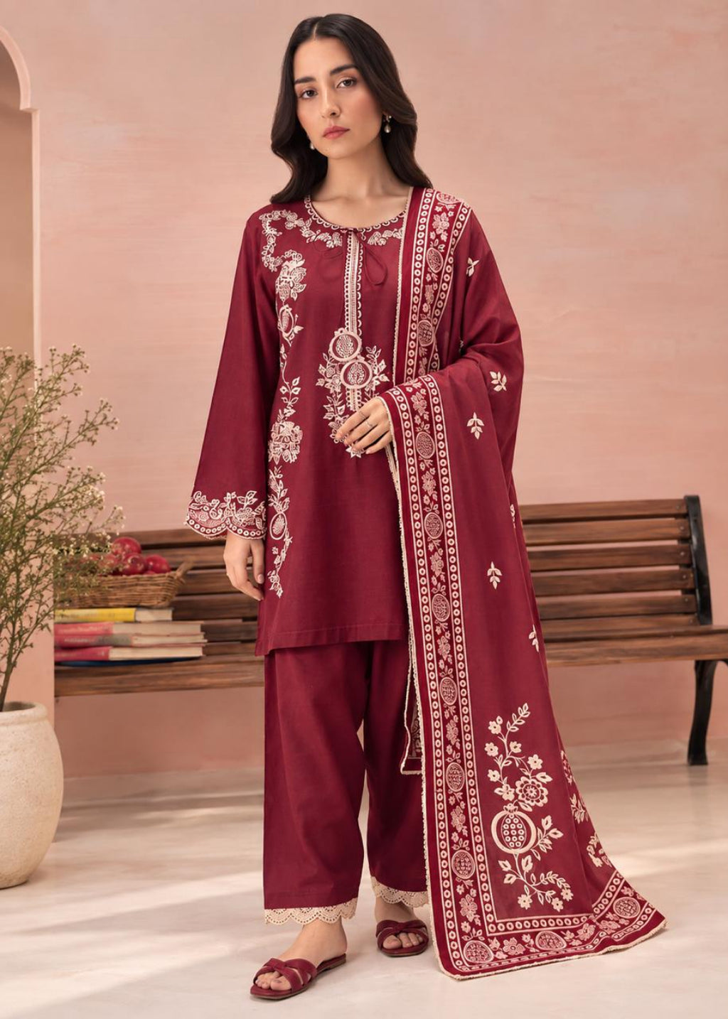 A woman models a three-piece maroon salwar kameez suit with white embroidery, featuring a long kurta, matching trousers, and a coordinating dupatta, set against a warm pink-beige background with a wooden bench.