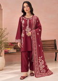 A woman models a three-piece maroon salwar kameez suit with white embroidery, featuring a long kurta, matching trousers, and a coordinating dupatta, set against a warm pink-beige background with a wooden bench.