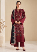 A woman models a traditional South Asian salwar kameez suit set in deep maroon and black, featuring intricate gold, blue, and white embroidery, set against a neutral beige background.