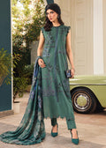 A woman models a green ethnic three-piece suit with matching dupatta, featuring intricate silver embroidery and printed patterns, standing in front of a vintage green car and a white building.