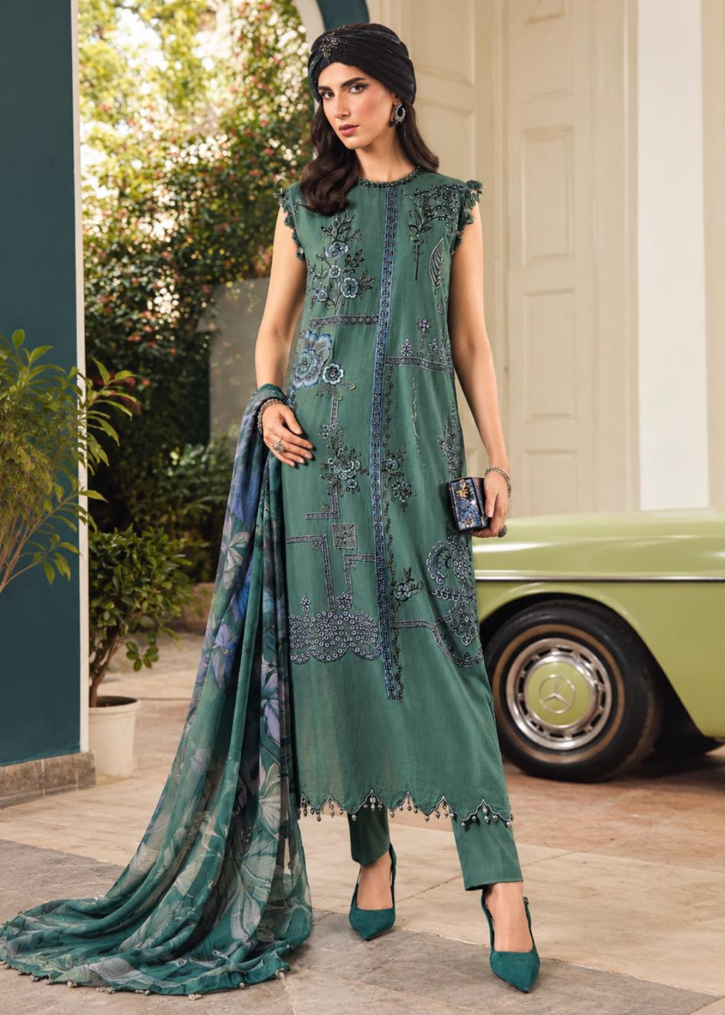 A woman models a green ethnic three-piece suit with matching dupatta, featuring intricate silver embroidery and printed patterns, standing in front of a vintage green car and a white building.