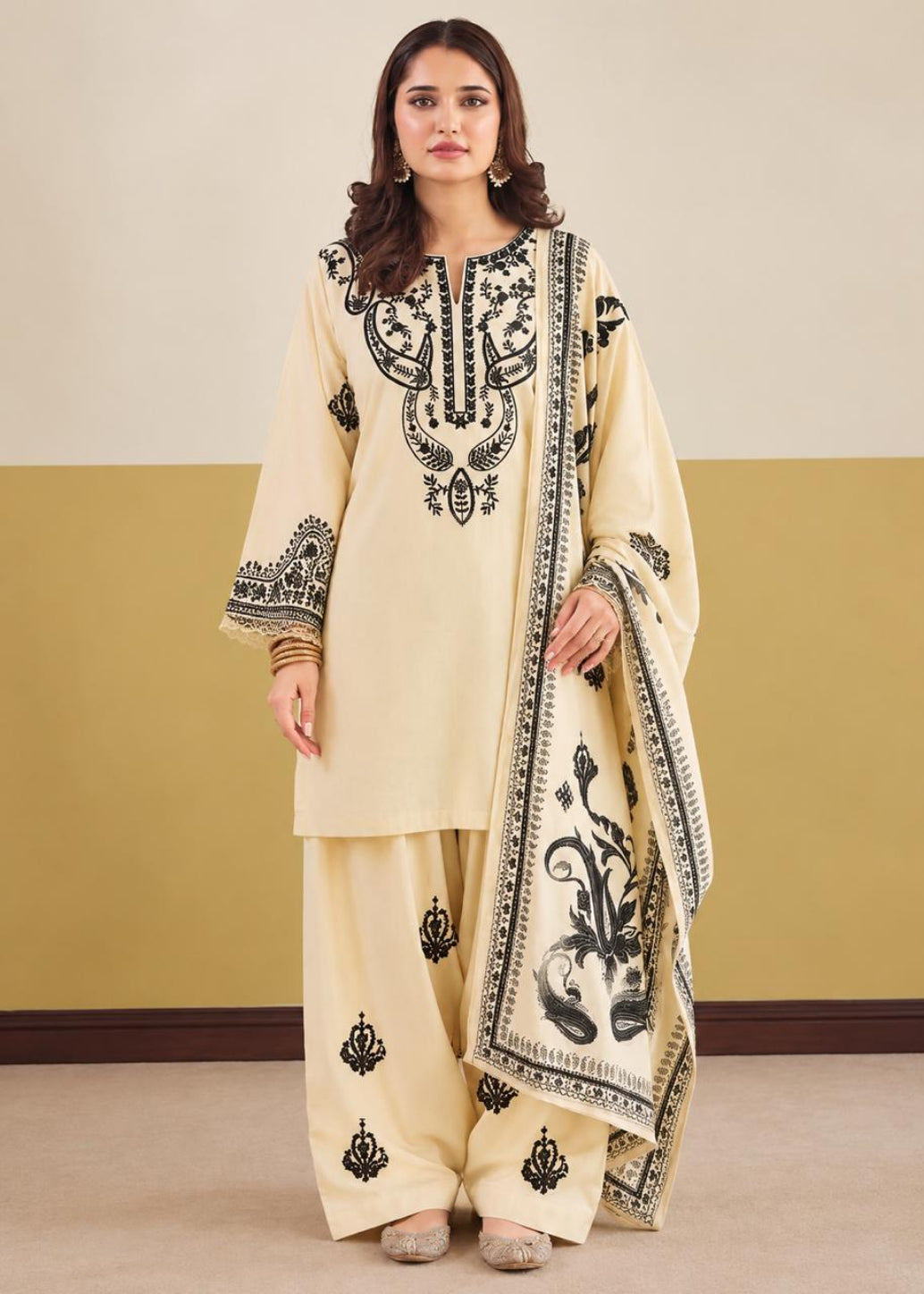 A woman modeling a cream-colored traditional South Asian outfit with black embroidery, featuring a long kurta, wide-leg pants, and a matching dupatta, set against a two-tone beige and mustard wall.