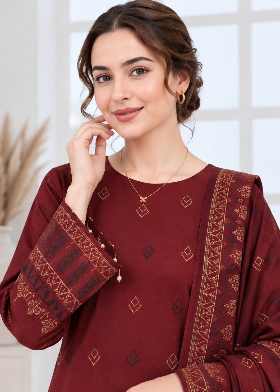 A woman wearing a deep maroon traditional kurta with gold geometric patterns and a matching dupatta, shown in a bright indoor setting.