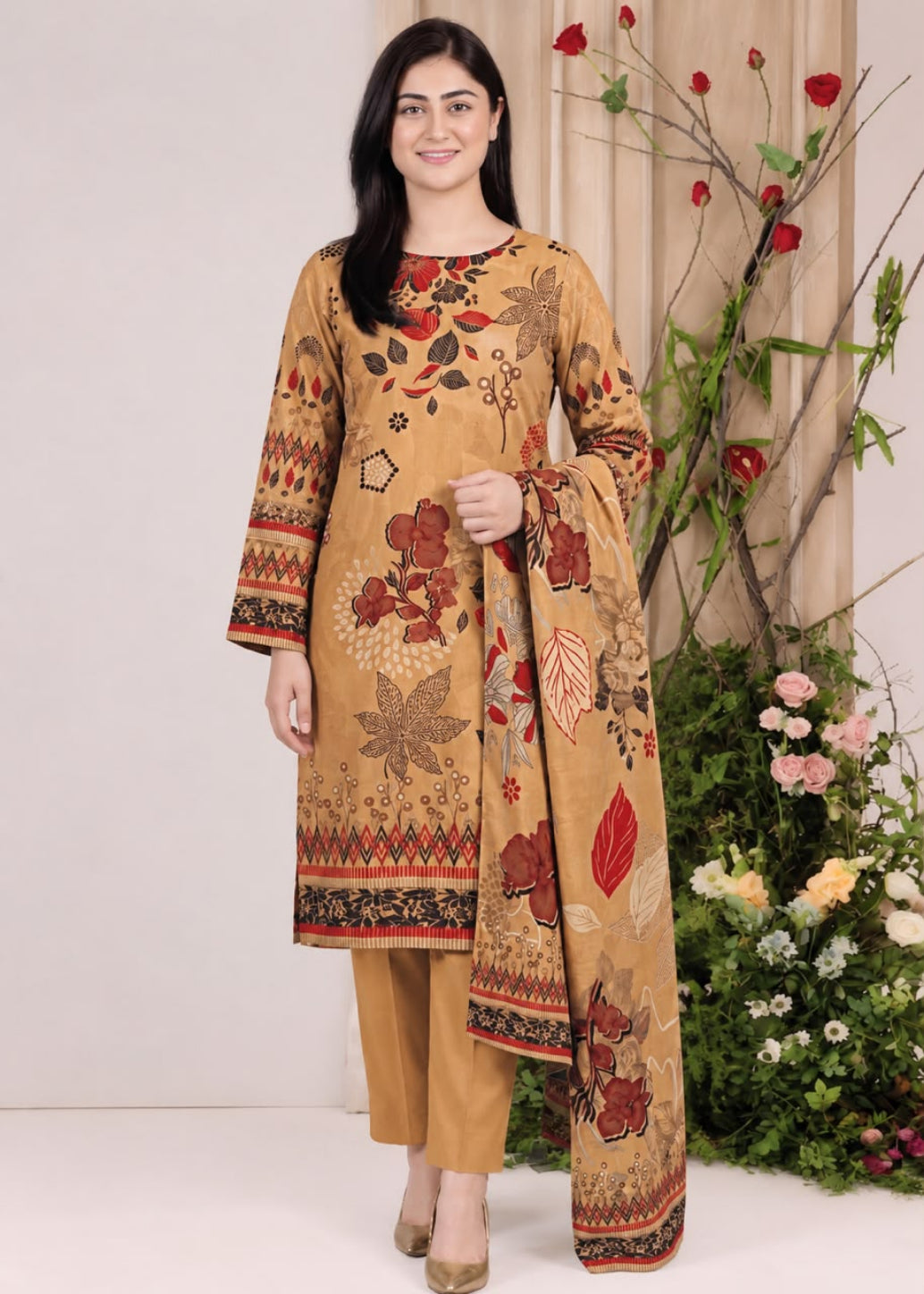A woman modeling a traditional three-piece salwar kameez suit in beige with a red, brown, and black floral print, standing in front of a neutral studio backdrop with decorative plants.