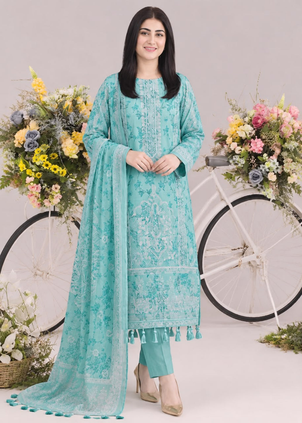 A woman modeling a traditional turquoise blue ethnic salwar kameez set with matching dupatta, standing in front of a white bicycle decorated with flowers against a light beige studio backdrop.