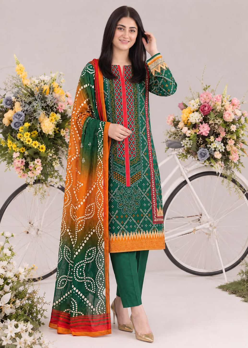 A woman models a traditional three-piece salwar kameez suit set. The kurta is forest green with red and gold geometric patterns, paired with matching green trousers and a dupatta featuring a green-to-orange gradient with white floral prints.