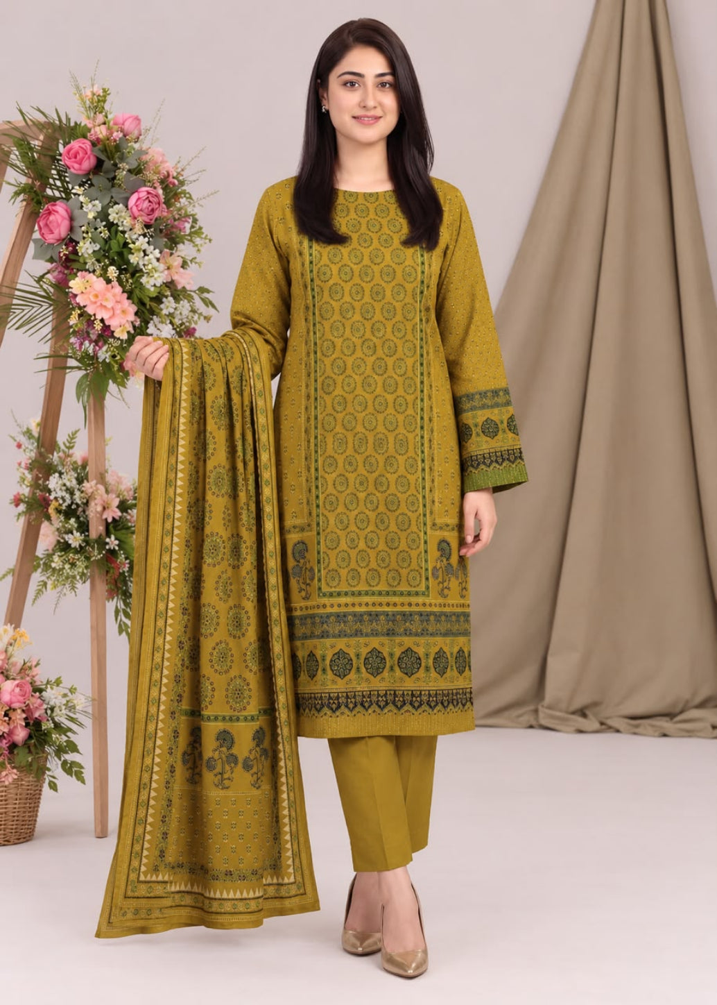 A woman models a three-piece olive green ethnic suit with intricate black and gold geometric patterns, consisting of a long kurta, matching trousers, and a dupatta, set against a neutral studio backdrop with floral decorations.