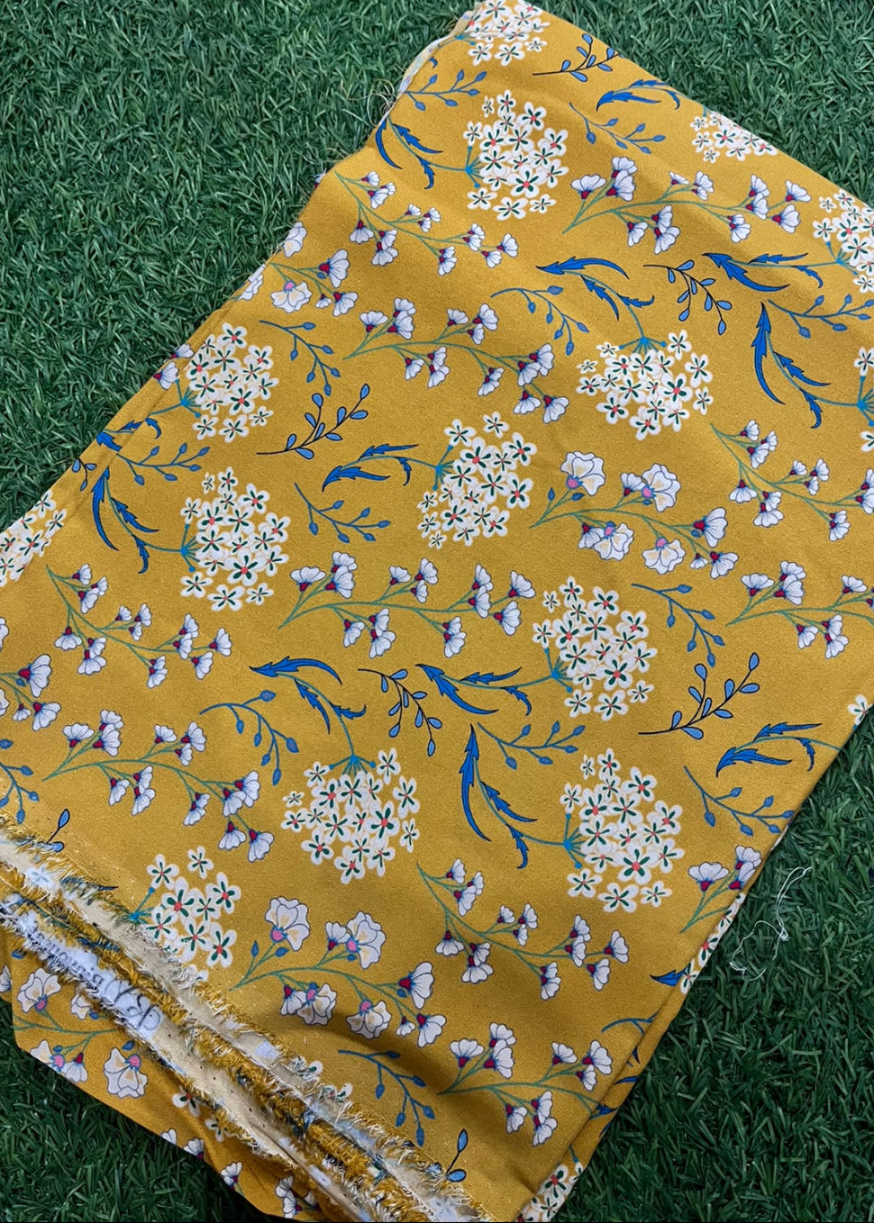 A folded piece of mustard yellow fabric with a blue and white floral print, laid flat on a green grass-like surface.