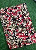 A folded piece of fabric with an intricate abstract pattern in red, black, beige, and brown, laid flat on a green grass-like surface.