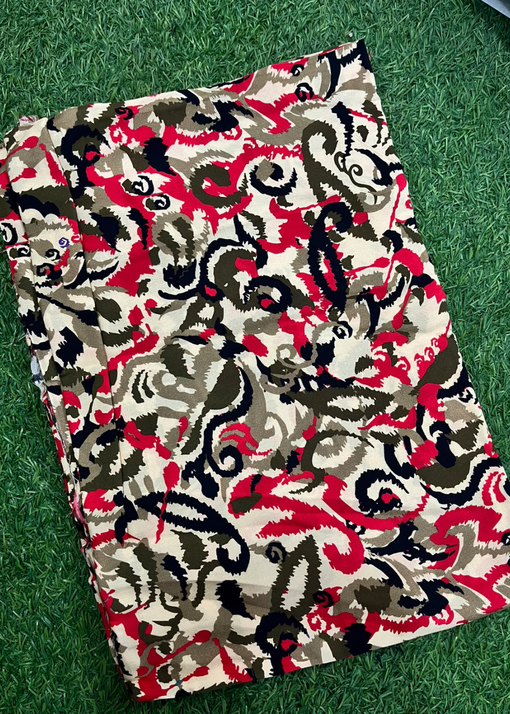 A folded piece of fabric with an intricate abstract pattern in red, black, beige, and brown, laid flat on a green grass-like surface.