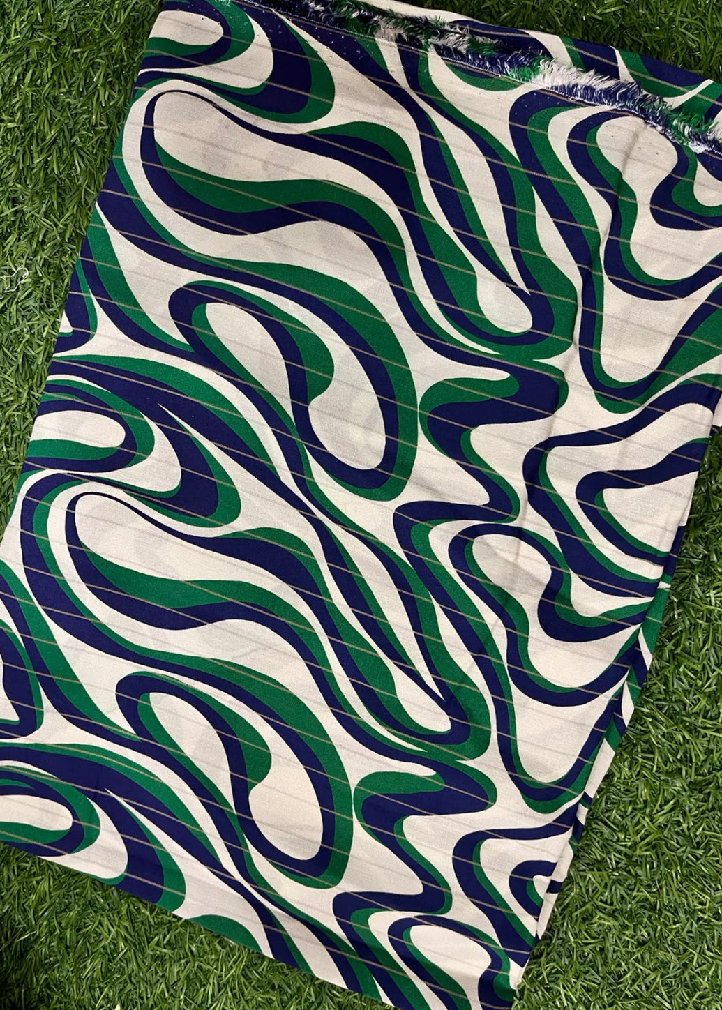 A square fabric item, likely a napkin, small tablecloth, or scarf, featuring a bold abstract wavy pattern in green, navy blue, and cream, laid flat on a green grass background.