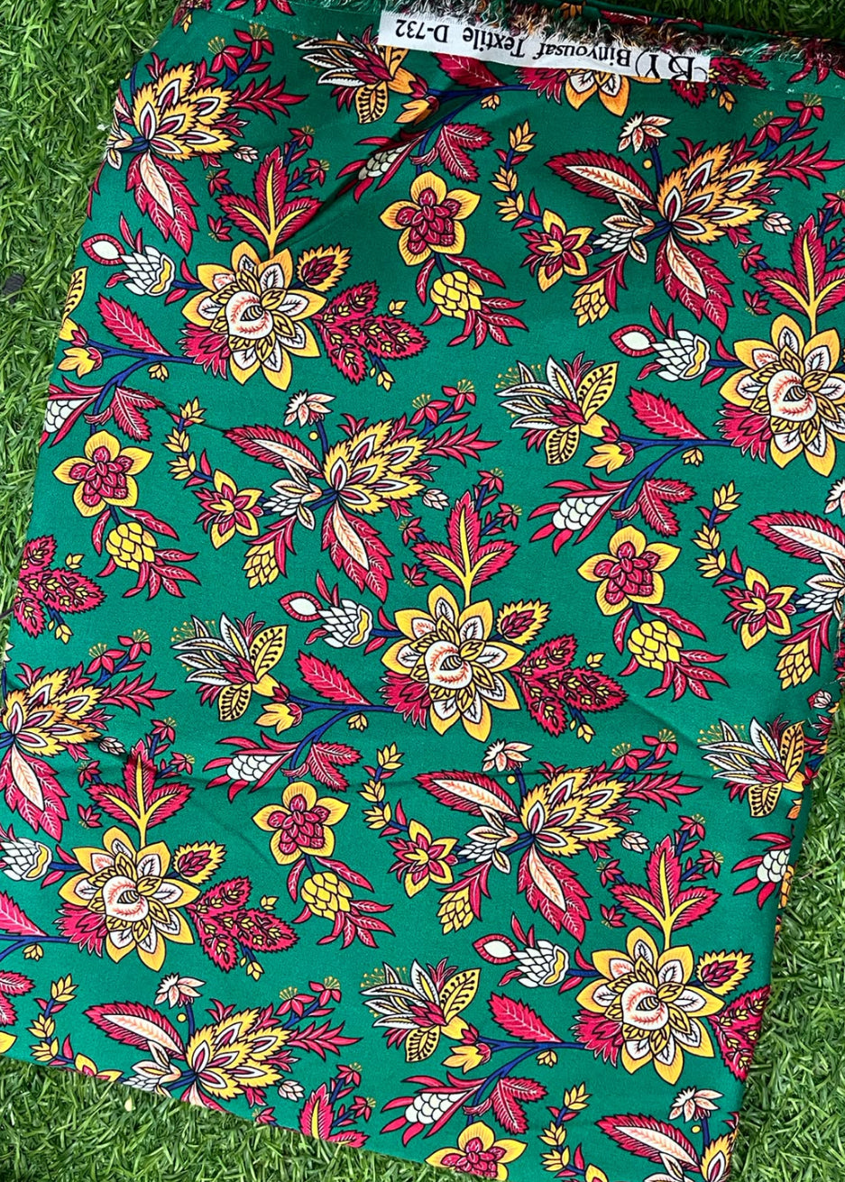 A folded piece of green fabric with a colorful floral block print pattern, laid flat on a green grass background. The print features yellow, pink, and red flowers with blue and brown details.