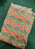 A square textile with a geometric diamond pattern in orange, olive green, yellow, and white, laid flat on a green grass-like surface.