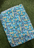 A folded textile item, likely a blanket or throw, featuring a colorful floral pattern in teal, navy, mustard yellow, and white, laid flat on a green grass-like surface.