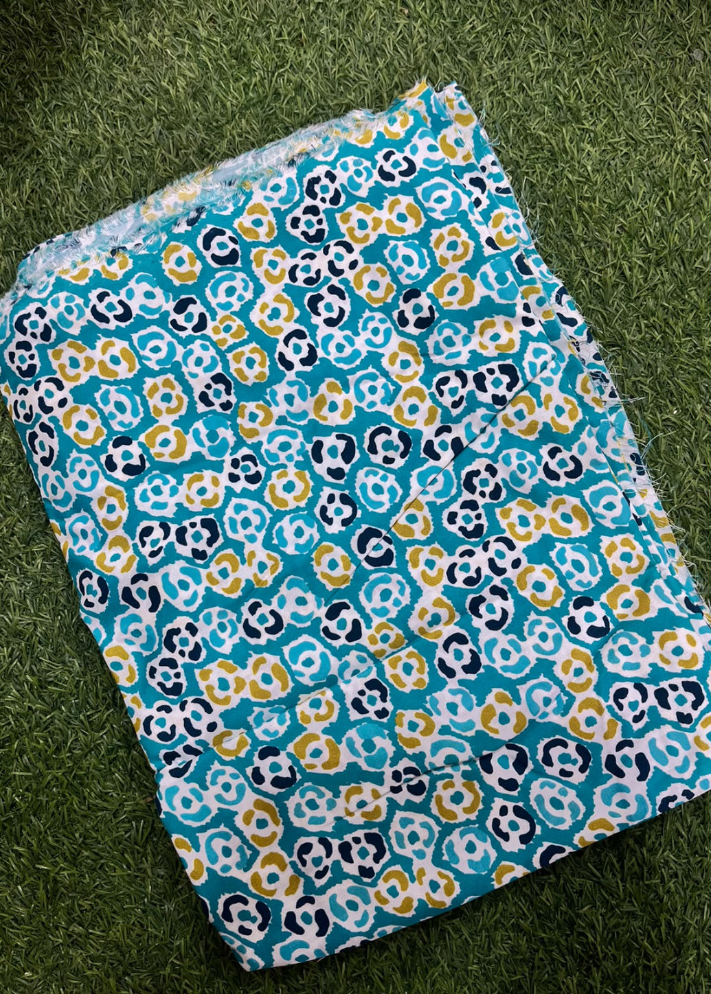 A folded textile item, likely a blanket or throw, featuring a colorful floral pattern in teal, navy, mustard yellow, and white, laid flat on a green grass-like surface.