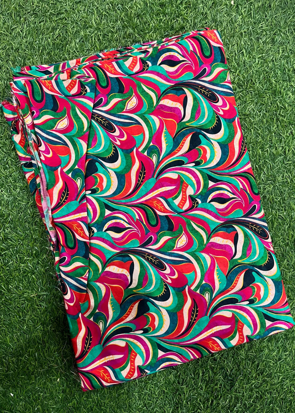A folded piece of fabric with a vibrant, multicolored abstract swirl pattern, laid flat on green grass.