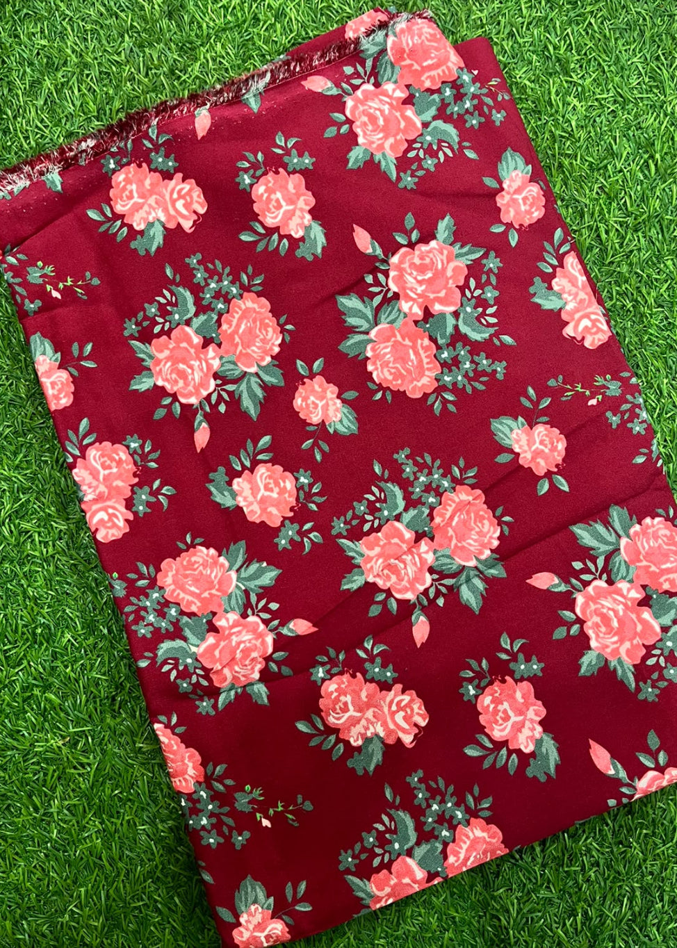 A folded piece of deep red fabric with a pink and green floral rose pattern, laid flat on a green grass background.
