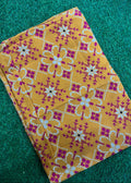 A folded piece of mustard yellow fabric with a magenta and white geometric pattern, laid flat on a green textured background.
