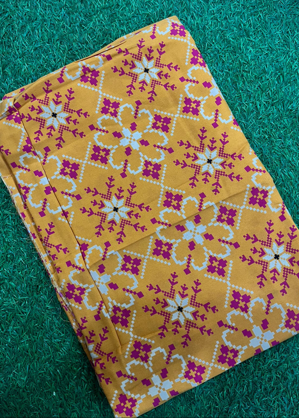 A folded piece of mustard yellow fabric with a magenta and white geometric pattern, laid flat on a green textured background.