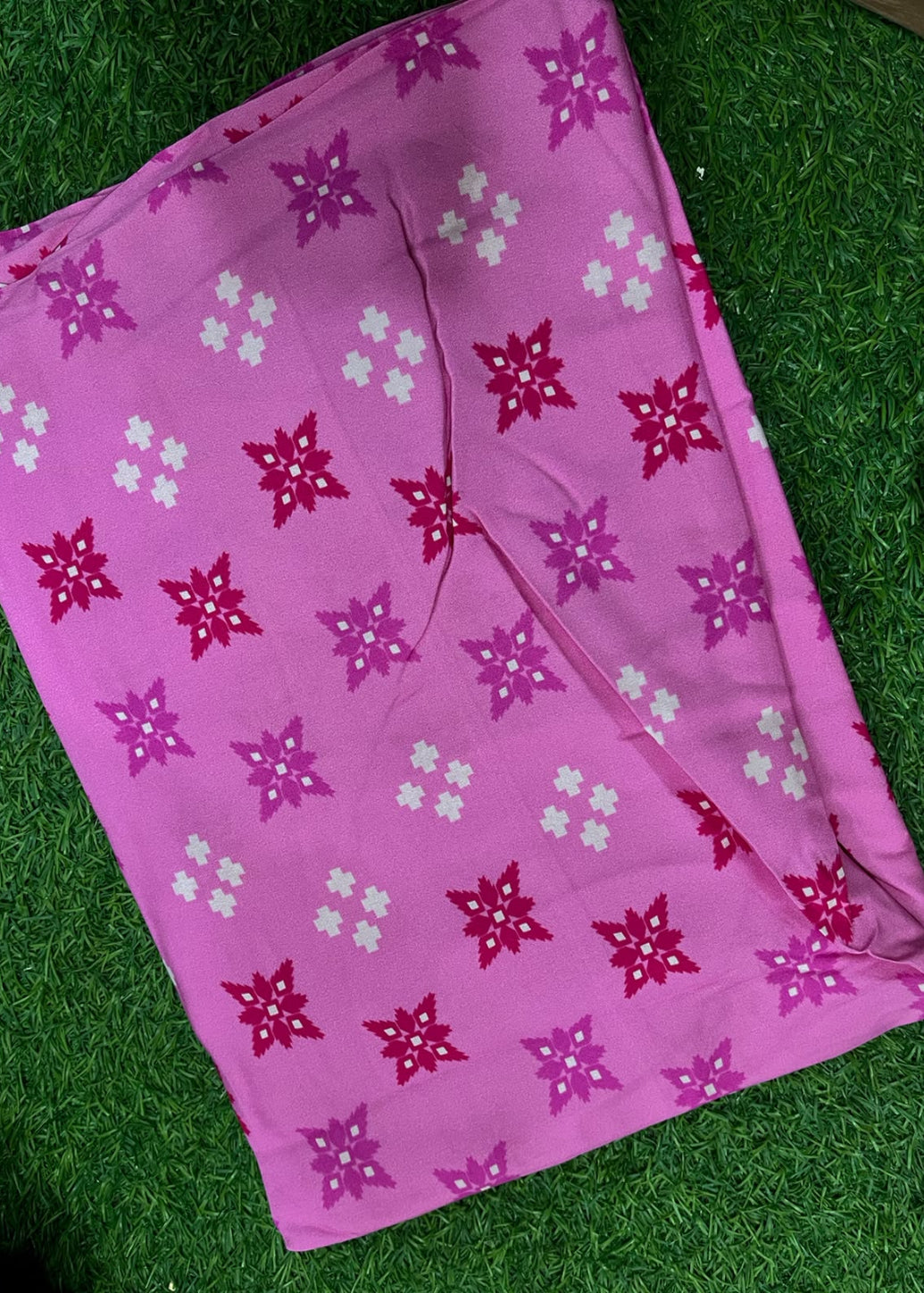 A pink fabric item, likely a scarf or sari, featuring a repeating pattern of magenta and white floral snowflake motifs, laid flat on a green artificial grass background.