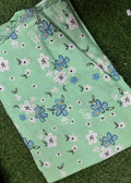 A mint green fabric item, likely a sheet or blanket, featuring a floral pattern of blue, pink, and white flowers with black outlines, laid flat on a green grass background.