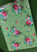 A light green fabric item, likely a scarf or bandana, featuring a vintage-style floral print with pink, red, blue, and white flowers with green leaves, laid flat on a green grass background.