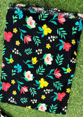A black fabric item, likely a skirt or scarf, featuring a colorful floral print with pink, yellow, and white flowers and green leaves, laid flat on green grass.