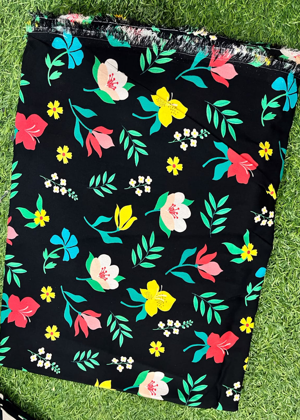 A black fabric item, likely a skirt or scarf, featuring a colorful floral print with pink, yellow, and white flowers and green leaves, laid flat on green grass.