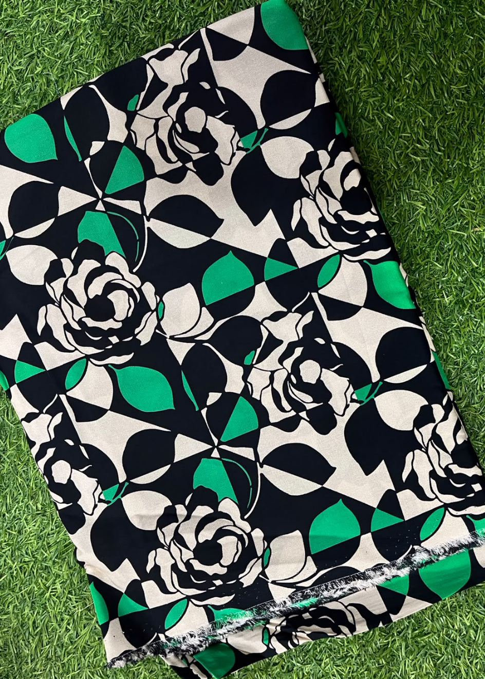 A folded piece of fabric with a bold geometric floral print in black, white, and green, laid flat on a green grass-like surface.