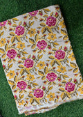 A folded piece of cream-colored fabric with a vibrant floral pattern featuring pink roses, yellow flowers, and green leaves, laid flat on a green grass background.