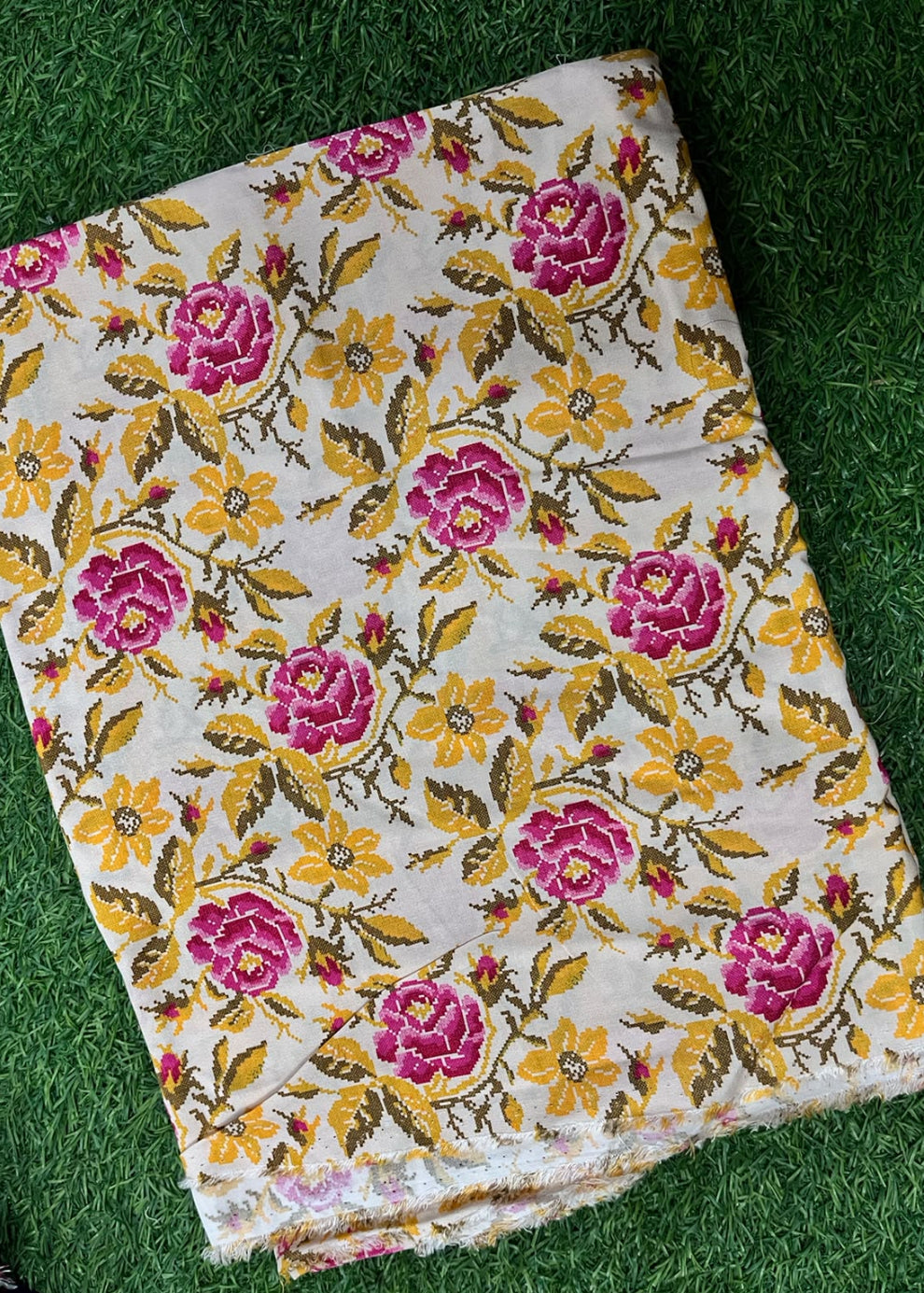 A folded piece of cream-colored fabric with a vibrant floral pattern featuring pink roses, yellow flowers, and green leaves, laid flat on a green grass background.
