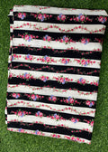 A folded fabric piece with horizontal black and white stripes, decorated with pink and purple floral garlands, laid flat on green grass.