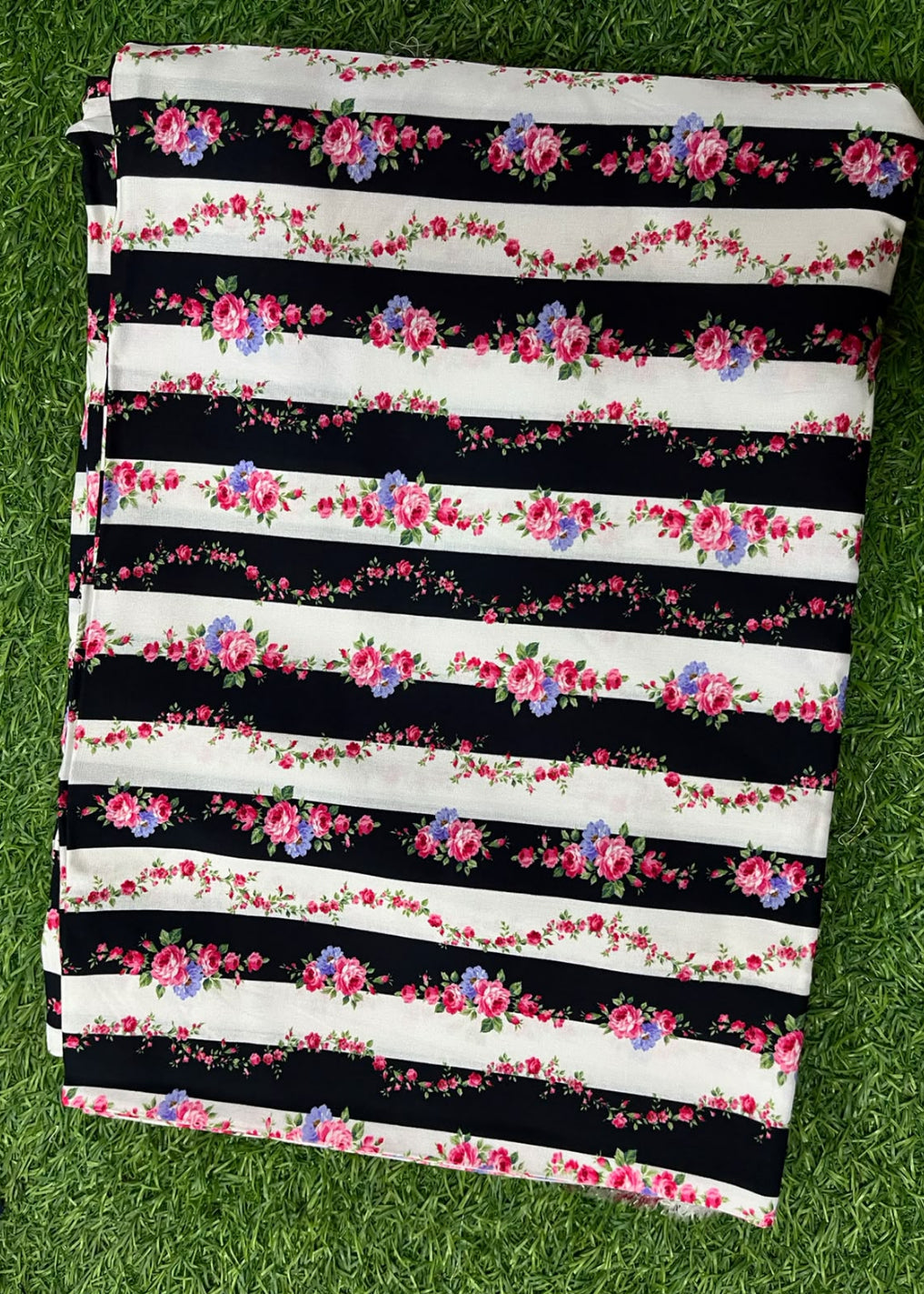 A folded fabric piece with horizontal black and white stripes, decorated with pink and purple floral garlands, laid flat on green grass.