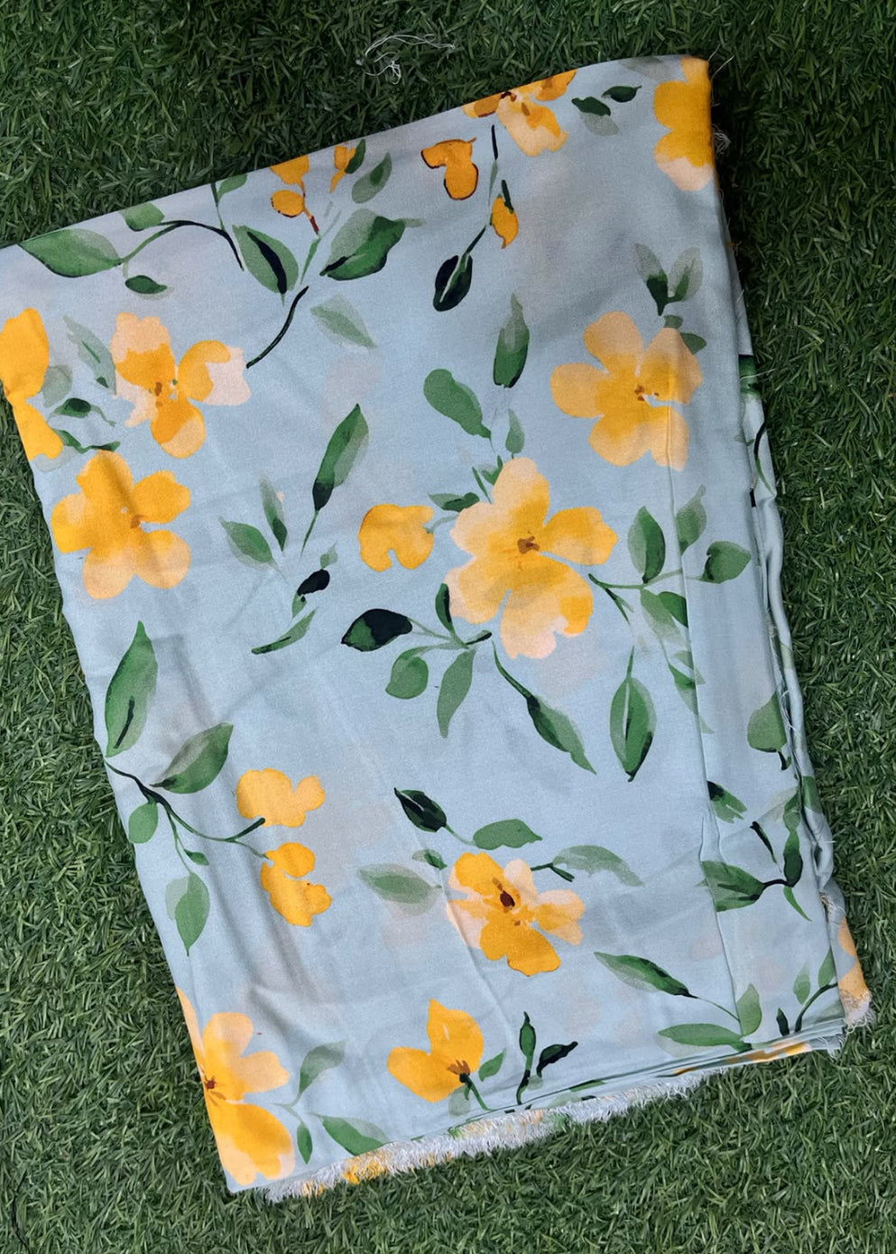 A folded piece of light blue fabric with a yellow and green floral print, laid flat on a green grass background.