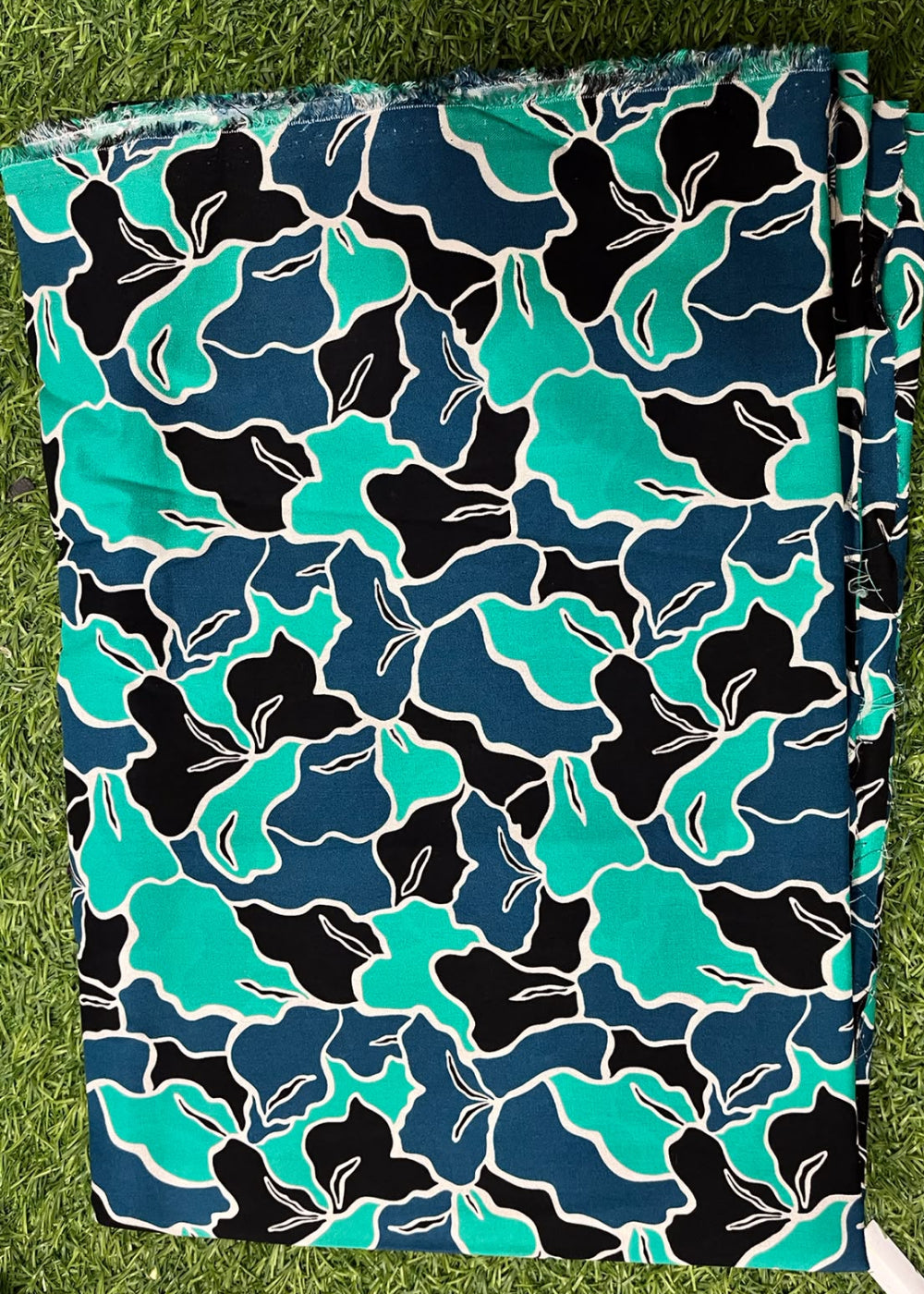 A folded piece of fabric with a bold abstract floral pattern in teal, navy, black, and white, laid flat on green grass.