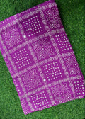 A purple bandhani fabric sheet with white dotted patterns, laid flat on a green grass background.