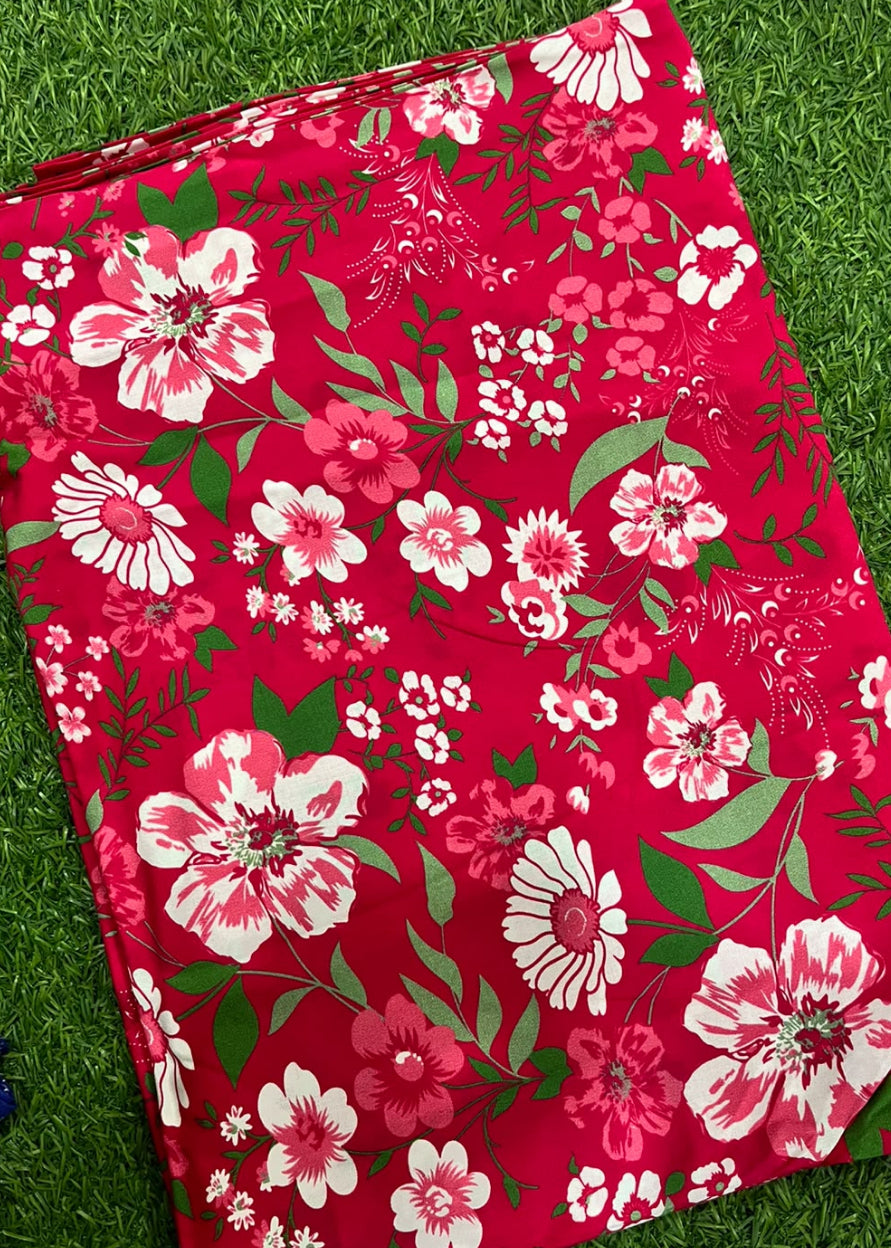 A rectangular red fabric item, likely a blanket or towel, featuring a vibrant floral print with pink and white flowers and green leaves, laid flat on green grass.