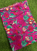 A folded piece of bright pink fabric with a bold floral print in green, orange, and white, laid flat on a green grass-like surface.