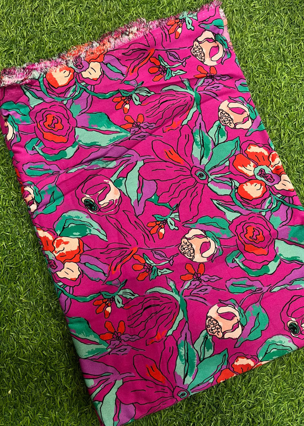 A folded piece of bright pink fabric with a bold floral print in green, orange, and white, laid flat on a green grass-like surface.