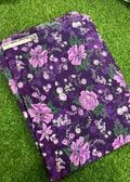A folded piece of purple floral fabric with pink, white, and green botanical patterns, laid flat on a green grass-like surface. A fabric label is visible at the top left edge.