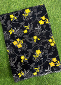 A black fabric item, likely a scarf or throw blanket, featuring a yellow and white floral vine pattern, laid flat on green grass.
