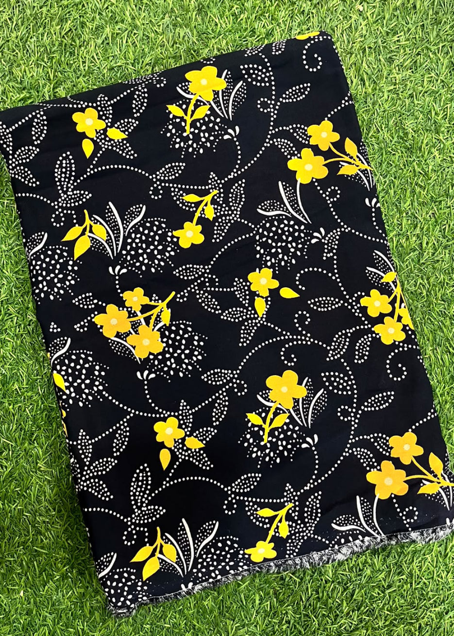A black fabric item, likely a scarf or throw blanket, featuring a yellow and white floral vine pattern, laid flat on green grass.