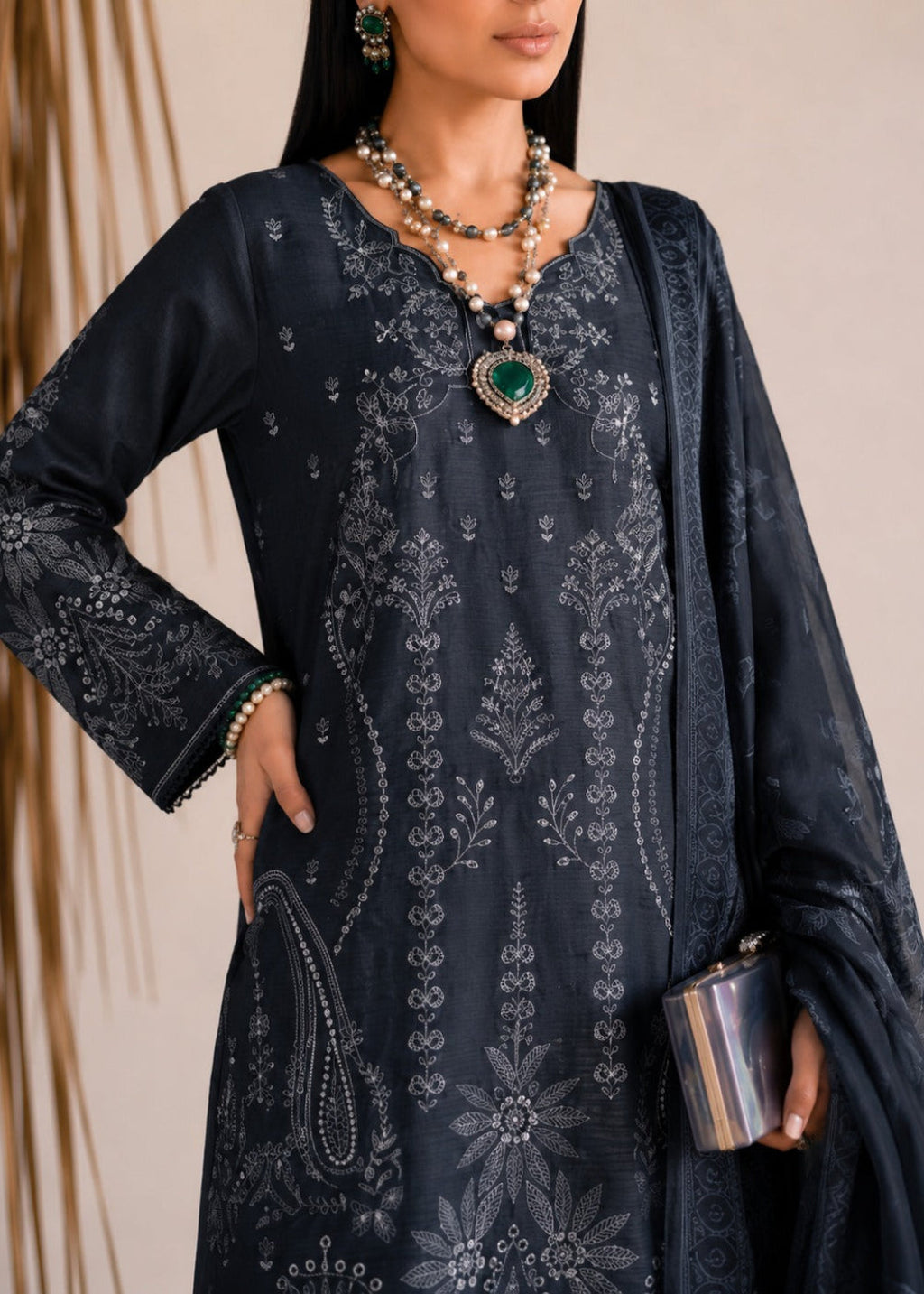 A woman models a dark navy blue traditional embroidered kurta set with matching dupatta and jewelry. The kurta features intricate silver and white floral embroidery, and the model is accessorized with a green heart pendant necklace and a silver clutch.