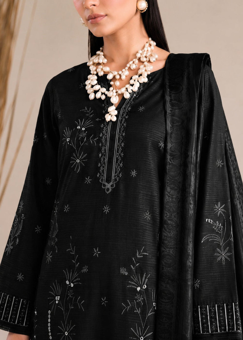 A close-up of a model wearing a black embroidered kurta with a matching dupatta and a statement pearl necklace, set against a neutral beige background.