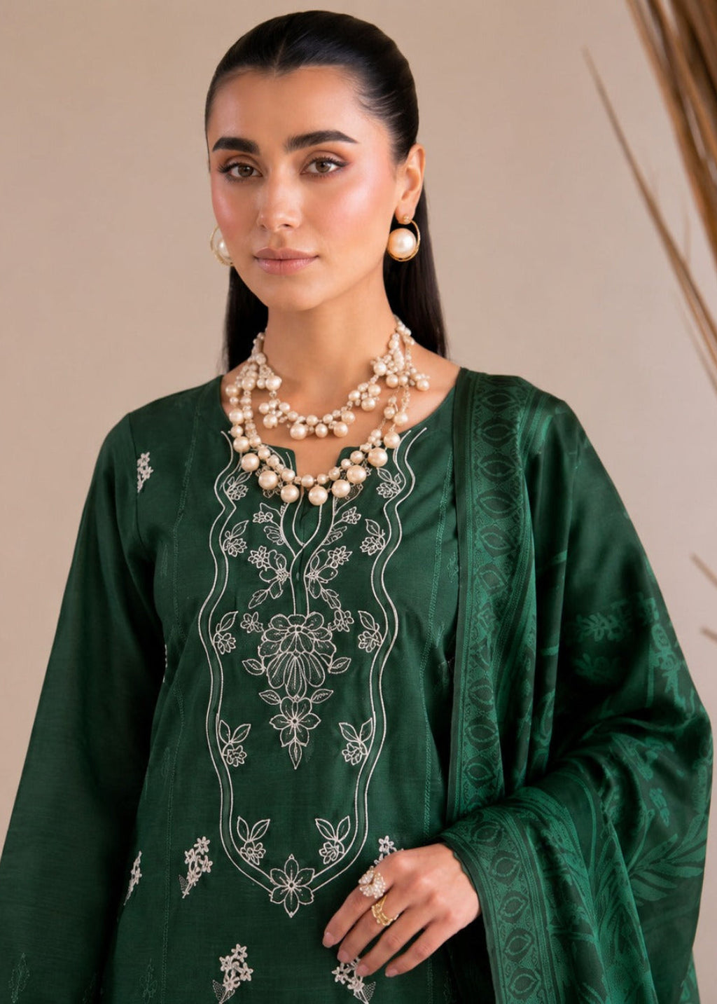 A woman modeling a dark green embroidered traditional outfit with white floral embroidery on the neckline and sleeves, accessorized with layered pearl necklaces and gold jewelry against a neutral beige background.