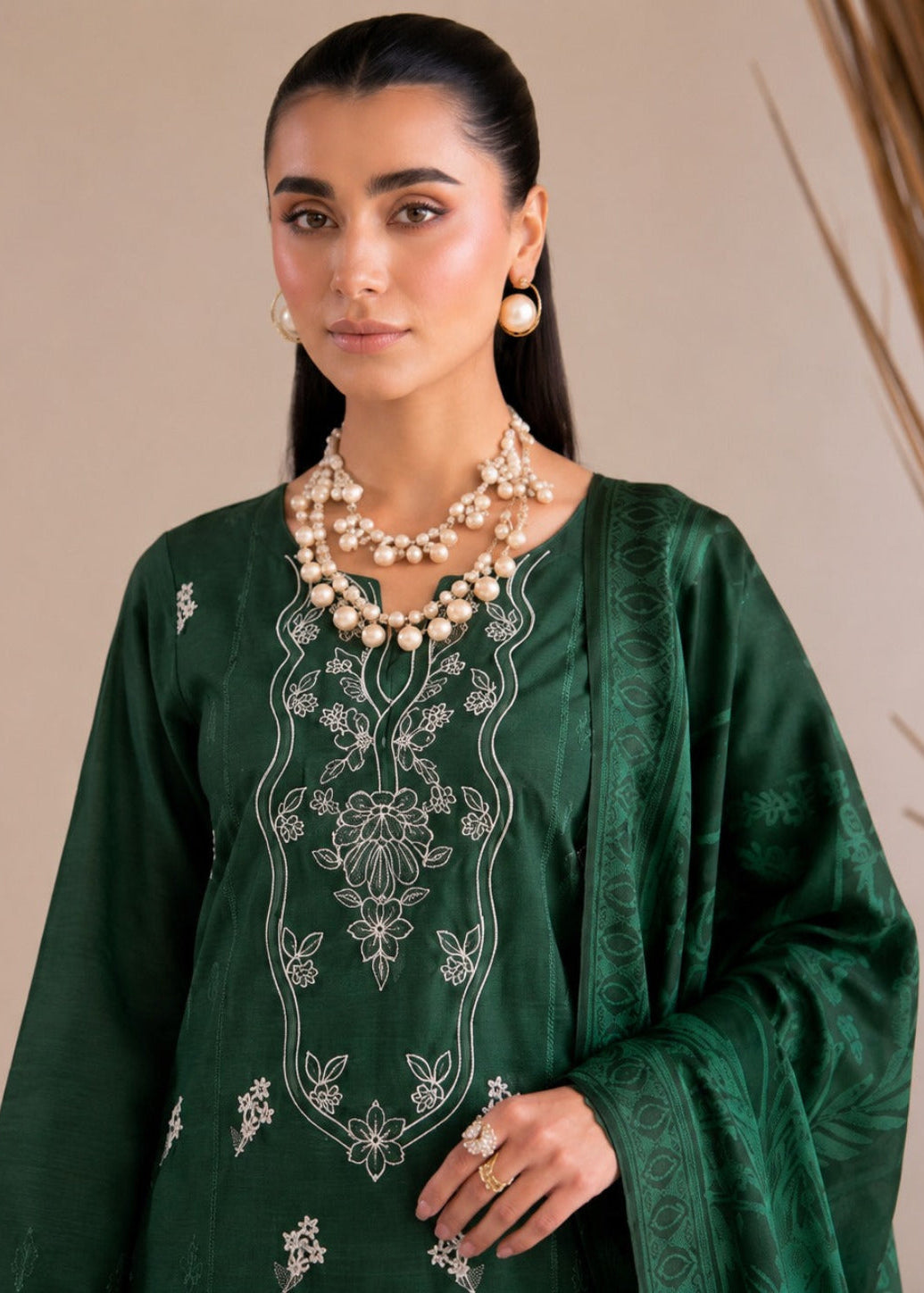 A woman modeling a dark green embroidered traditional outfit with white floral embroidery on the neckline and sleeves, accessorized with layered pearl necklaces and gold jewelry against a neutral beige background.