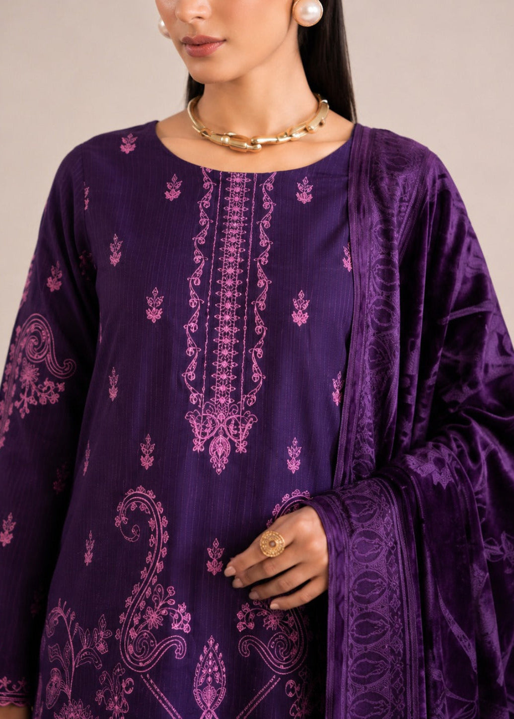 A woman wearing a deep purple traditional outfit with intricate magenta embroidery and a matching draped scarf, styled with gold jewelry against a softly blurred indoor background.
