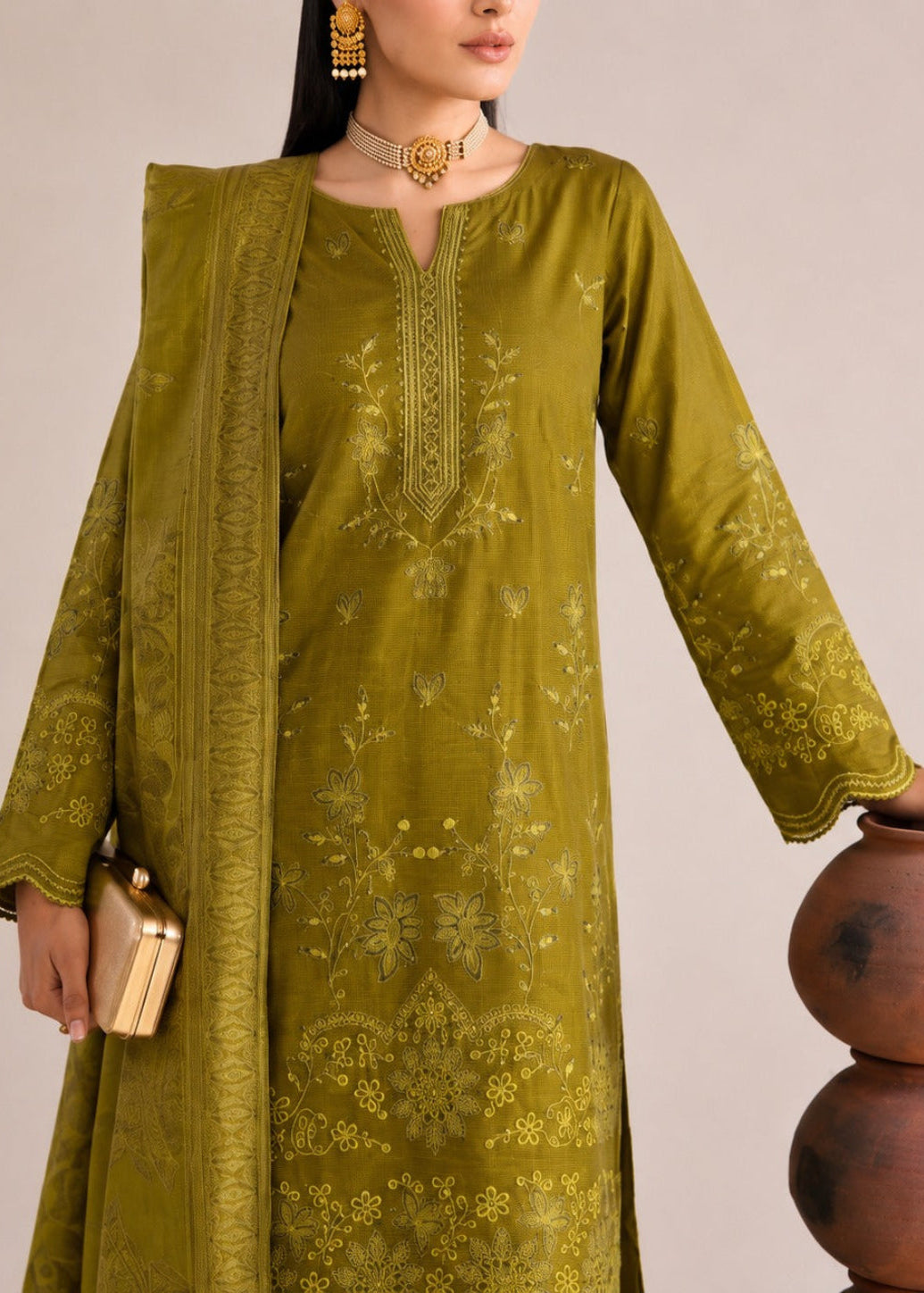A woman models an olive green embroidered kurta set with a matching dupatta and gold clutch, standing next to a terracotta pot against a neutral beige background.