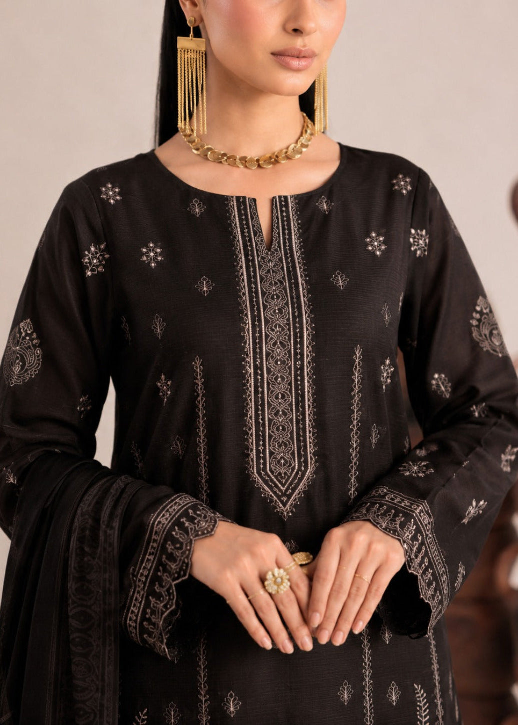 A woman wearing a black embroidered traditional outfit with metallic gold-toned jewelry, shown from the shoulders to the waist against a softly blurred indoor background.